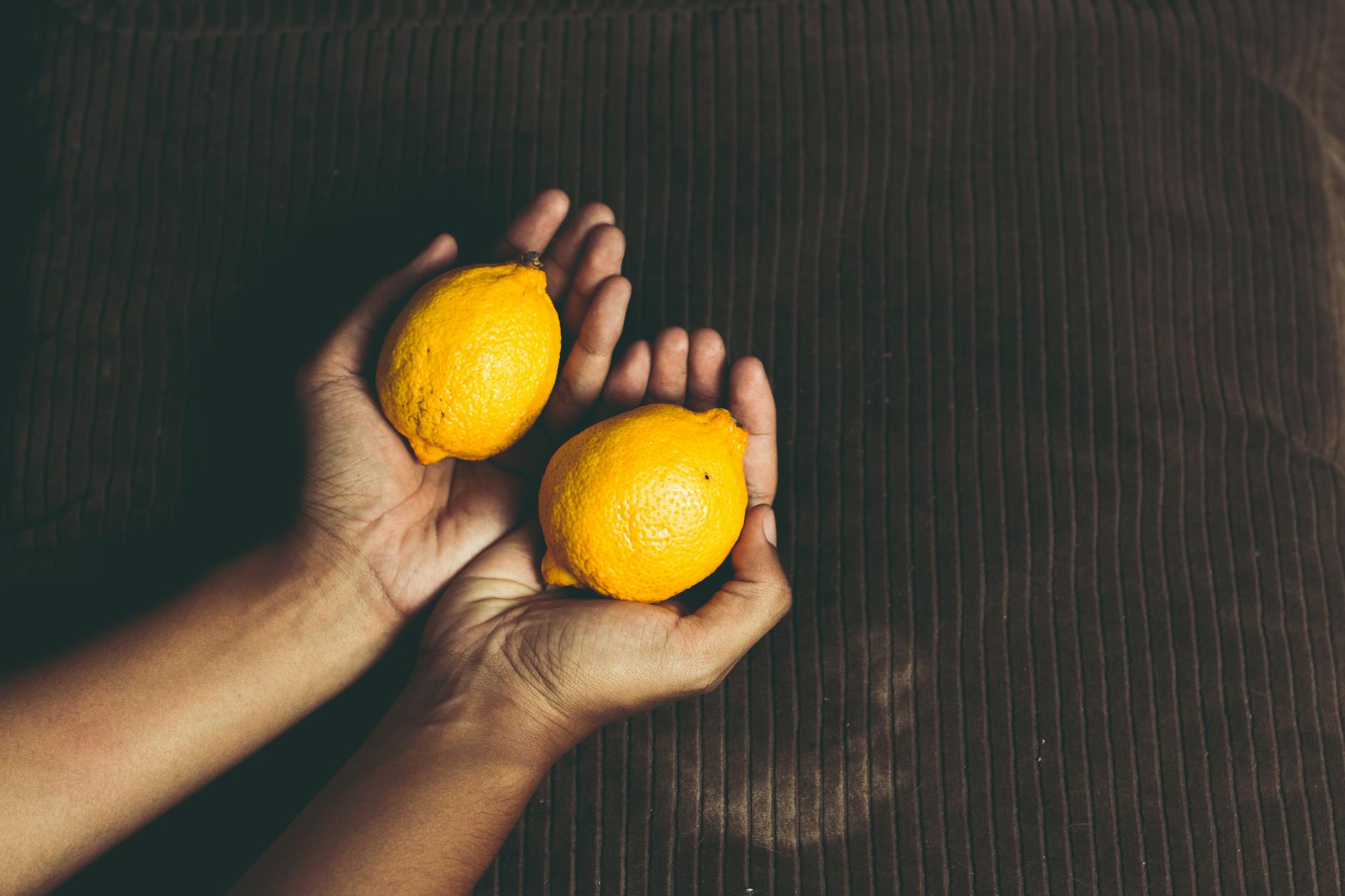 Close-up of two fresh lemons held in cupped hands on a brown surface, symbolizing freshness and natural sensation.