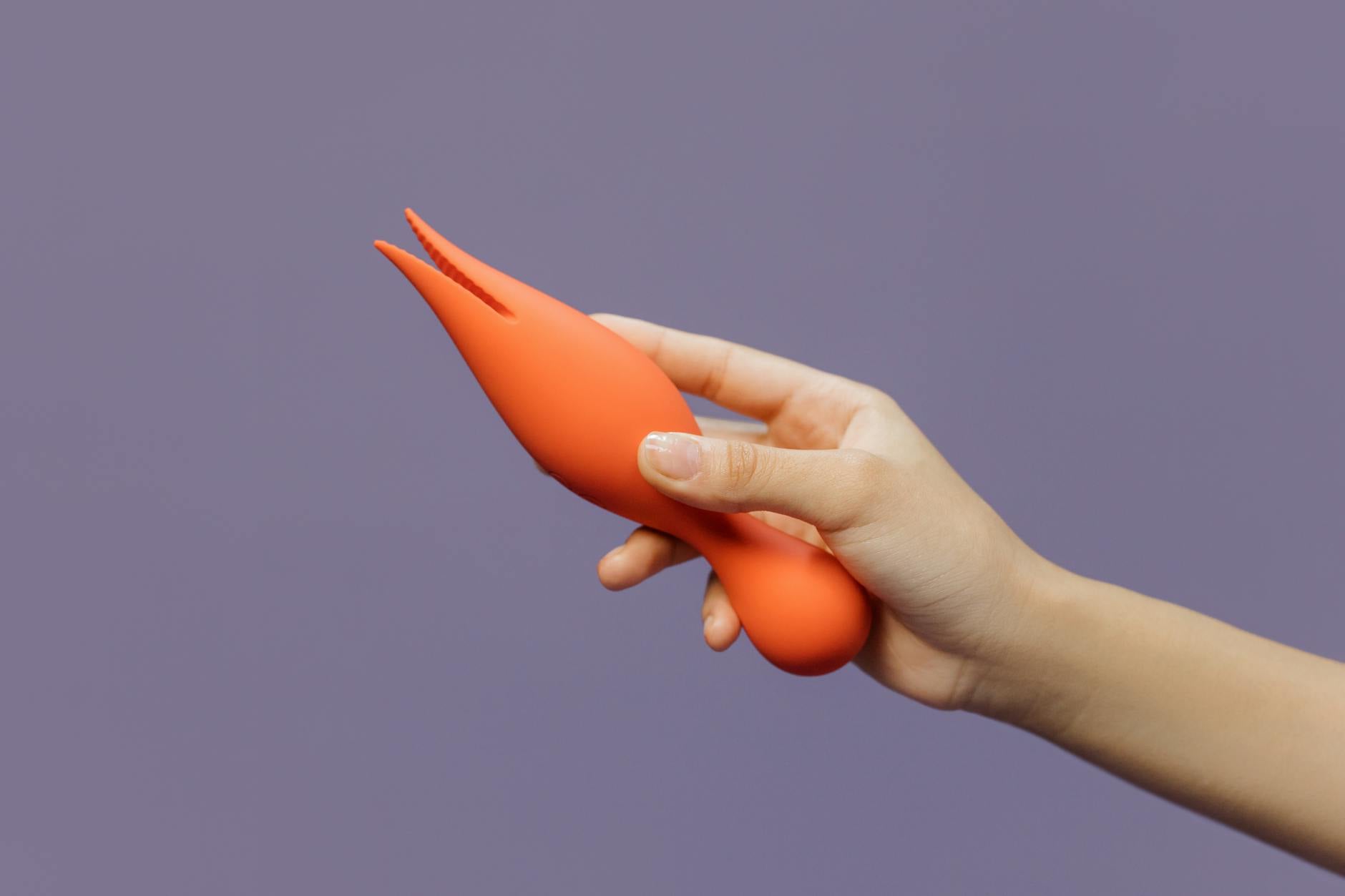 Close-up of a hand holding an orange vibrator against a purple minimalistic backdrop, representing modern intimate exploration